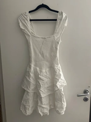 Vit volangklänning med smock - Supersöt vit kortklänning med smockad överdel och breda axelband. Kjolen har flera lager volanger som ger en luftig och lekfull känsla. Perfekt för varma dagar och sommarens alla tillfällen.