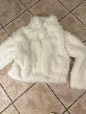 Vit kort pälsjacka med hög krage - Säljer en fluffig vit pälsjacka i kort modell med hög krage och långa ärmar. Jackan har en clean look utan synliga knappar eller dragkedja och är perfekt för att höja din vinterstil. Materialet är mjukt fuskpäls som känns lyxigt och trendigt.