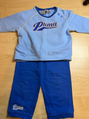 Blått set från Puma med tryck - Säljer ett blått tvådelat set från Puma med långärmad tröja och byxor. Tröjan har tryckknappar vid axeln och ett stort Puma-tryck framtill. Byxorna är i matchande blå färg med Puma-logga vid benet. Mjukt material, perfekt för en avslappnad stil.