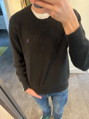 Svart tröja från Polo Ralph Lauren - Klassisk svart tröja från Polo Ralph Lauren med lila broderad logga på bröstet. Tröjan har rund halsringning, ribbade muddar och är tillverkad i mjukt material. Perfekt för en clean och stilren look.