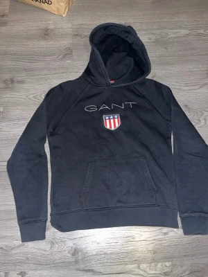 Svart hoodie från GANT med logga - Snygg svart hoodie från GANT med stor vit logga och ett broderat sköldmärke med stjärnor och ränder på bröstet. Klassisk känguruficka framtill och huva. Perfekt för en avslappnad och stilren look. Storlek 13-14 years old/158-164 cm som motsvarar XS, Hör av dig vid frågor. Mvh Edvin
