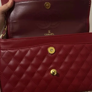 Röd Classic Flap handväska - Säljer en ikonisk röd Classic Flap handväska i quiltat läder med guldfärgade metalldetaljer och kedjerem. Väskan har dubbla lock, CC-logga framtill och elegant design som verkligen sticker ut. Perfekt för dig som vill ha något lyxigt och tidlöst.