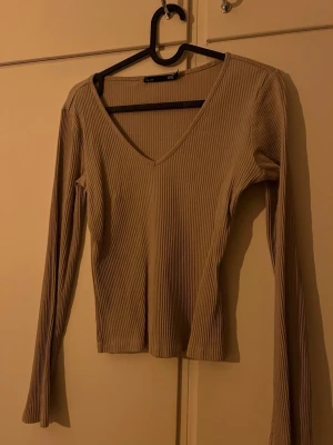 Beige ribbad långärmad topp - Trendig beige långärmad topp med v-ringning och ribbad struktur. Perfekt basplagg som är lätt att styla till olika outfits. Tillverkad i mjukt material för en bekväm känsla och har en smickrande passform.