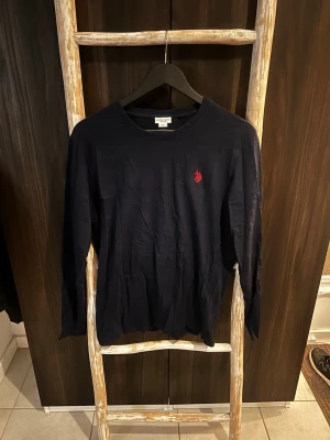 Mörkblå långärmad tröja U.S. Polo Assn. - Mörkblå långärmad tröja från U.S. Polo Assn. med rund halsringning och röd broderad logga på bröstet. Tröjan är i mjuk bomull och har en klassisk, avslappnad passform. Perfekt för en stilren och enkel look.