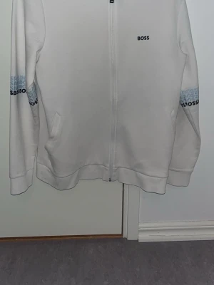 Vit zip hoodie från BOSS - Snygg vit hoodie från BOSS med dragkedja framtill och diskret logga på bröstet. Ärmarna har blå detaljer med BOSS-tryck. Två fickor på sidorna och ribbade muddar. Perfekt för en clean och stilren look.