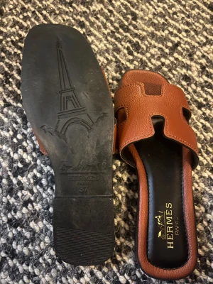 Bruna Hrmès Oran sandaler i läder - Snygga bruna Oran sandaler från Hrmès i äkta läder med ikonisk H-design på ovandelen. Svart innersula med guldfärgad logga och präglad Eiffeltornet-detalj under sulan. Perfekta för en trendig och lyxig look.