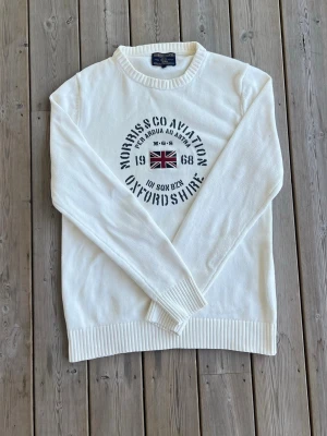 Vit stickad tröja med tryck - Snygg vit stickad tröja med rund hals och ribbade muddar. Framtill finns ett tryck med brittisk flagga och texten 'Morriss Co Aviation Oxfordshire 1968'. Perfekt till dig som gillar klassisk stil med en twist.