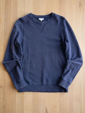Mörkblå sweatshirt med rund hals från Sunspel - En stilren mörkblå sweatshirt med rund halsringning och ribbade muddar vid ärmslut och nederkant. Tröjan har en klassisk relaxed fit och ett diskret X-detalj vid halsen. Perfekt för en avslappnad look och enkel att matcha med jeans eller joggers.