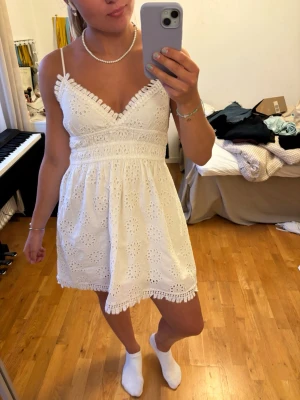 Vit spetsklänning med smala axelband - Söt vit kortklänning med broderade blommor och spetsdetaljer. Klänningen har smala axelband, v-ringning och volangkant vid urringningen samt nederkanten. Perfekt för sommarens alla tillfällen.