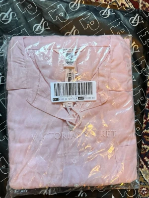 Ljusrosa pyjamas från Victoria's Secret - Supermysig ljusrosa pyjamas från Victoria's Secret med klassisk skjortkrage och knappar framtill. Setet består av långärmad överdel och byxa med resår i midjan. Materialet är mjukt och har en subtil glans, perfekt för chillkvällar hemma.