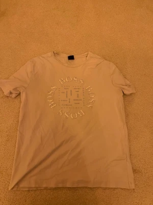Vit BOSS t-shirt med tryck - Snygg beige t-shirt från BOSS med rund hals och stort tryck framtill där det står BOSS i cirkel och namn på olika städer. Klassisk passform och korta ärmar. Perfekt till jeans eller shorts för en clean och stilren look.