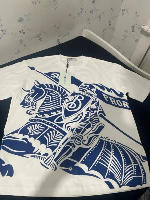 Vit t-shirt med blått riddarmotiv - Säljer en vit t-shirt med stort blått tryck av en riddare på häst och texten 'PRORSUM' framtill. T-shirten har klassisk passform och korta ärmar. Perfekt för dig som gillar unika och grafiska prints.