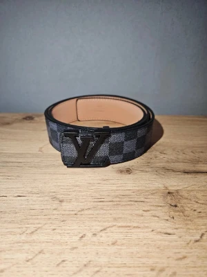 Louis Vuitton rutigt bälte i skinn - Snyggt bälte från Louis Vuitton med klassiskt rutigt mönster i svart och grått. Bältet har en stilren LV-spänne i svart metall och insida i ljust skinn. Perfekt accessoar för att lyfta din outfit.