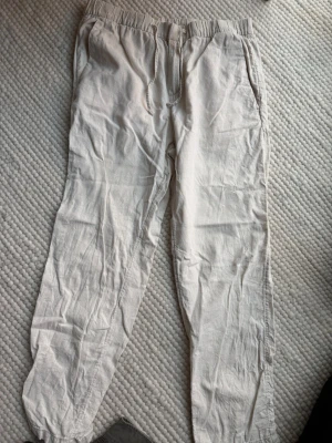 Beiga linnebyxor från H&M - Snygga beiga linnebyxor från H&M i regular fit. Byxorna har resår i midjan, sidofickor och dragsko med knappdetalj. Perfekta för en avslappnad och stilren look. Tillverkade i ett lätt och luftigt linnematerial.
