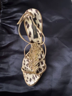Roberto Cavalli skor! Guldiga pumps med leopardmönster - Unika pumps från Roberto Cavalli med leopardmönstrad innersula och tunna guldiga remmar. Dekorerade med ett stort guldfärgat blad med glittrande stenar framtill. Smal klack och öppen tå ger en elegant och trendig look. Obs: skon är enbart buren en gång, det fattas ett par kristaller men går att sätta dit nya lätt! Använd en endaste gång! 
