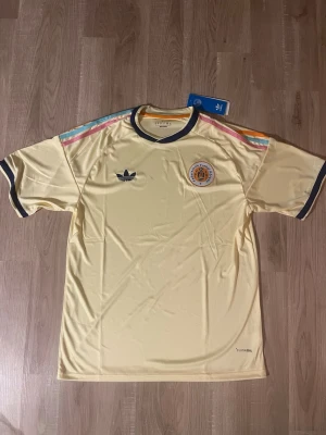 Curacao VM-2026 Fotbollströja - Snygg gul Curacao tröja VM-2026. Tröjan har färgglada ränder på axlarna i rosa, blått och orange samt mörkblå detaljer vid krage och ärmslut. Broderad logga och klubbmärke på bröstet. Tillverkad i lätt och ventilerande material. HELT NY!