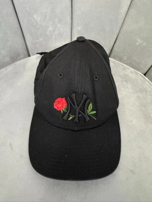 Svart keps med broderad ros och NY-logga - Snygg svart keps med böjd skärm och broderad NY-logga framtill. Dekorerad med en röd ros och gröna blad. Justerbart spänne baktill för perfekt passform. Tillverkad i bomullstyg och passar till streetwear och casual outfits.