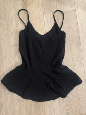 Svart peplum linnetopp - Snygg svart linnetopp med smala axelband och peplumskärning. Toppen har en v-ringad halsringning och är tillverkad i ett mjukt tyg som ger en skön känsla. Perfekt att styla med jeans eller kjol för en trendig look.
