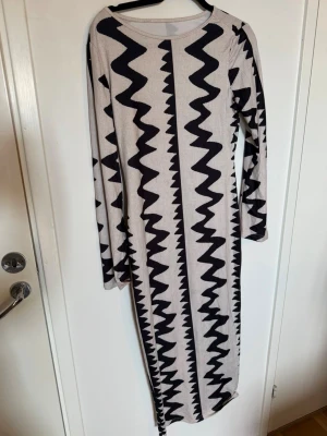 Långärmad mönstrad långklänning - Snygg långklänning med svartvitt zigzag-mönster. Klänningen har lång ärm och rund halsringning, figurnära passform och är i ett stretchigt material. Perfekt för dig som vill sticka ut med en cool grafisk look.