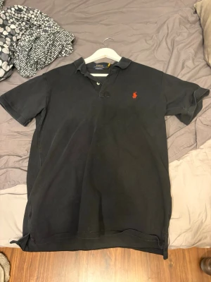 Svart pikétröja från Polo Ralph Lauren - Klassisk svart pikétröja från Polo Ralph Lauren med korta ärmar och krage. Tröjan har två knappar vid halsen och den ikoniska röda logotypen broderad på bröstet. Perfekt till jeans eller shorts för en clean och stilren look.