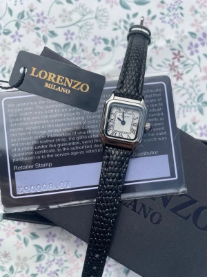 Lorenzo Milano svart klocka - Stilren klocka från Lorenzo Milano med fyrkantig silverfärgad boett och vit urtavla med svarta romerska siffror. Svart läderband med textur ger en lyxig känsla. Perfekt accessoar för dig som vill ha något klassiskt men ändå modernt. Pris kan diskuteras och mer information finns