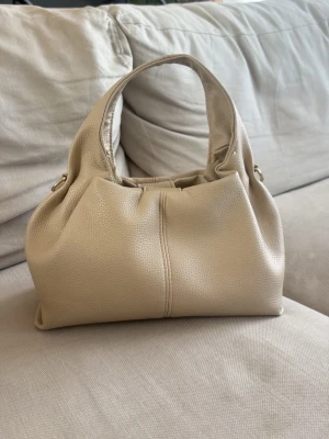 Beige handväska  - Stilren beige handväska i mjuk skinnimitation med rundade former och korta handtag. Väskan har en enkel men trendig design med guldfärgade metalldetaljer på sidorna. Perfekt för dig som gillar minimalistisk och modern stil.