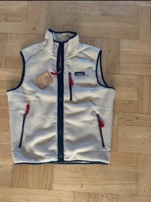 Beige fleeceväst från Patagonia - Säljer en krämvit fleeceväst från Patagonia med marinblå detaljer och dragkedjor med röda dragare. Västen har hög krage, tre fickor med dragkedja och är tillverkad i mjukt teddyfleece-material. Perfekt för lager-på-lager och outdoor-stil.