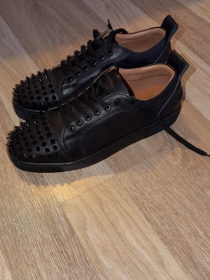 loubs Svarta läder nya  - Säljer ett par svarta sneakers i läder med coola svarta nitar på tån. Skorna har svarta skosnören, svarta öglor och en klassisk låg siluett. Insidan är beige och materialet känns lyxigt. Perfekta för dig som vill sticka ut med din stil. Helt nya strl 42-43