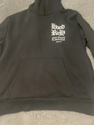 Svart hoodie med Hoodrich-tryck - Svart hoodie med stor vit grafisk text på ryggen och mindre tryck på bröstet. Klassisk känguruficka framtill och huva. Materialet ser ut att vara mjuk bomullsblandning, perfekt för en streetwear-look, och byxorna kommer med båda storlek M