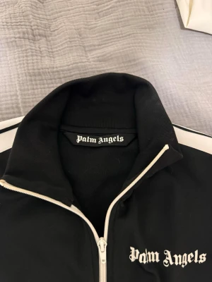 Palm Angels svart track jacket - Svart track jacket från Palm Angels med vita ränder längs ärmarna och vit dragkedja. Klassisk hög krage och logga på bröstet. Perfekt för en streetwear-look och riktigt snygg till jeans eller sweatpants.