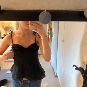Svart zara topp - Säljer en svart linnetopp med tunna axelband som knyts på axlarna. Toppen har en lätt utsvängd nederdel och söt rynkdetalj vid bysten. Materialet är tunt och ger en luftig känsla, perfekt för varma dagar.