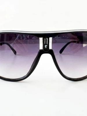 Svarta Carrera pilotbågar solglasögon - Snygga solglasögon från Carrera med svart båge och stora pilotformade glas. Glasen är tonade i grått och ger en cool retrovibe. Perfekta för dig som vill sticka ut med en klassisk men trendig accessoar.