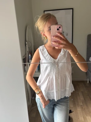 Vit ärmlös blus med spetsdetaljer - Söt vit blus i bomull med spetskant och broderade detaljer. Blusen är ärmlös, har v-ringning och knappar framtill. Perfekt till jeans eller kjol för en fräsch och somrig look. Toppen har prislappen kvar!