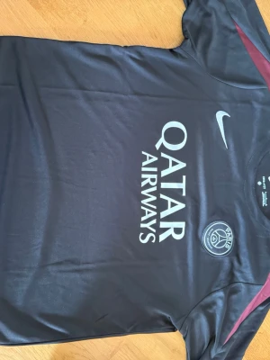 PSG svart matchställ från Nike - Säljer ett svart PSG matchställ från Nike med Qatar Airways-tryck på tröjan och klubbmärke på både tröja och shorts. Setet har vinröda detaljer på axlar och sidor, samt vita Nike-loggor. Tillverkat i lätt och snabbtorkande material, perfekt för fotbollsträning.