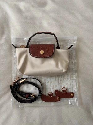 Beige och brun minihandväska - Snygg minihandväska i beige med bruna detaljer och guldfärgad knapp. Väskan har både handtag och avtagbar axelrem samt två små läderdetaljer. Perfekt för dig som gillar stilrena accessoarer med klassisk design.