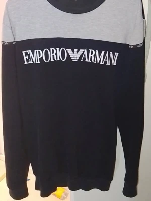 Emporio Armani svart och grå tröja - Snygg långärmad tröja från Emporio Armani i svart med grå axelparti och kontrasterande vit logga över bröstet. Tröjan har rund halsringning och mjukt material, perfekt för en stilren och avslappnad look.