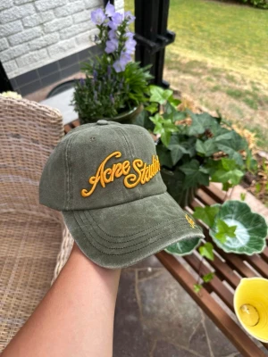 Olivgrön keps från Acne Studios - Säljer en olivgrön keps från Acne Studios med gul broderad logga framtill och på skärmen. Klassisk böjd skärm och justerbar rem bak. Tillverkad i mjuk bomull med en avslappnad passform. Perfekt accessoar för en cool och trendig look.