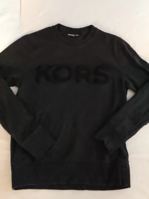 Svart tröja från Michael Kors - Snygg svart långärmad tröja från Michael Kors med stor sammetsliknande KORS-logga framtill. Tröjan har rund halsringning och ribbade muddar vid ärmslut och nederkant. Perfekt för en stilren och avslappnad look.