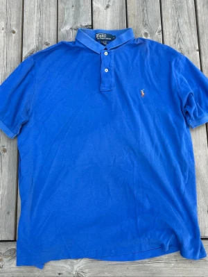 Blå pikétröja från Polo Ralph Lauren - Klassisk blå pikétröja från Polo Ralph Lauren med korta ärmar och krage. Tröjan har två knappar framtill och den ikoniska broderade loggan på bröstet. Perfekt till jeans eller shorts för en avslappnad men snygg look.