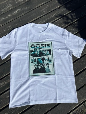 Vit Oasis t-shirt med tryck - Säljer en vit t-shirt med stort tryck av bandet Oasis på framsidan. T-shirten har klassisk rund hals och korta ärmar. Perfekt för dig som gillar bandtröjor och vill ha en stilren look med ikoniskt motiv.