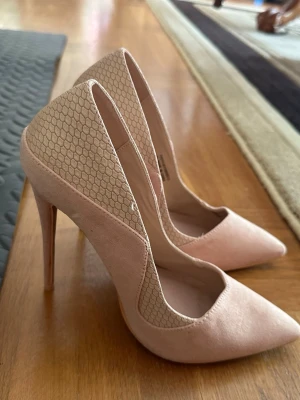 Beiga pumps med mönstrad detalj - Säljer ett par snygga pumps i beige mocka med spetsig tå och höga stilettklackar. Baksidan har en cool hexagonmönstrad textil 