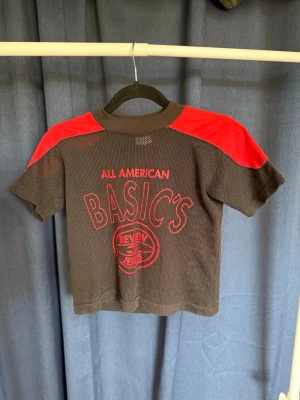 Svart och röd mesh t-shirt BASIC'S - Cool svart mesh t-shirt med röda axelpartier och trycket 'ALL AMERICAN BASIC'S REVIEW JEANS' framtill. Tillverkad i 60% bomull och 40% polyester. Perfekt för en avslappnad och sportig stil.