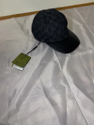Svart keps med GG-mönster - Snygg svart keps med GG-mönster över hela tyget och böjd skärm i svart läder. Kepsen har justerbar rem bak och är tillverkad i en mix av textil och läder. Perfekt för dig som vill ha en exklusiv och trendig accessoar. GG keps 