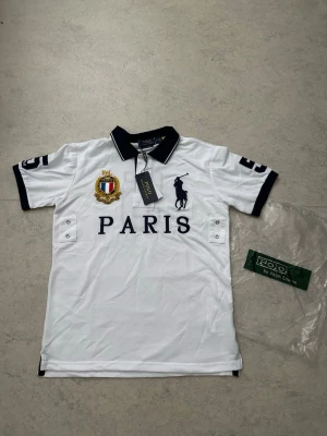 Vit Polo Ralph Lauren Paris t-shirt - Säljer en helt ny Polo Ralph Lauren Paris Big Pony pikétröja i storlek S.  Svart pikétröja med Big Pony-logga och Paris-brodyr på bröstet. Helt ny, oanvänd och etiketter kvar.  Information:  * Märke: Polo Ralph Lauren * Modell: Paris Big Pony * Färg: Svart * Storlek: S * Helt ny med etiketter  Skickas eller hämtas enligt överenskommelse.