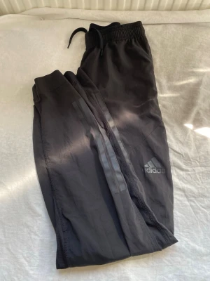  Adidas mjukisbyxor - Svarta mjukisbyxor från Adidas med diskreta svarta ränder längs benen och klassisk logga på ena benet. Byxorna har resår i midjan med dragsko och är tillverkade i ett lätt och bekvämt material. Perfekta för chill eller träning.Pris kan diskuteras 