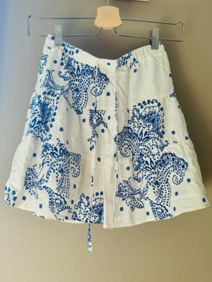 Vit kortkjol med blått blommönster - Söt vit kortkjol med blått blommigt mönster och volangdetaljer. Kjolen har en avslappnad passform och knytband i midjan. Perfekt för varma dagar och ger en fräsch sommarkänsla.