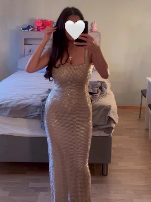Petra Maxi Dress Gold - Babyboo - Säljer en glittrig guldig långklänning med smala axelband och figurnära passform. Klänningen har ett skimrande material som fångar ljuset och ger en lyxig känsla. Perfekt för dig som vill sticka ut och glänsa på nästa event. (I sista bilden kan ni se en liten skada längst ner o längst bak på klänningen SOM EJ SYNS men de kan lösas o så kommer även extra pärlor med) Använd en gång! 