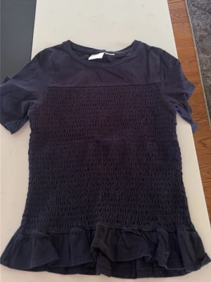 Mörkblå topp med volangdetalj - Snygg mörkblå topp med korta ärmar och rund hals. Toppen har smockad mittdel och volang längst ner för en extra touch. Perfekt till jeans eller kjol för en trendig look. Storlek 146/152 