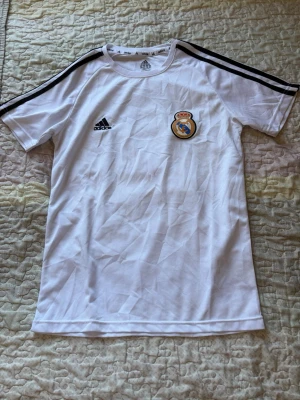 Vit Real Madrid tröja från Adidas - Säljer en vit Real Madrid t-shirt från Adidas med klubbens emblem på bröstet och svarta ränder längs axlarna. Tröjan har rund hals och är tillverkad i ett lätt och ventilerande material, perfekt för träning eller match.
