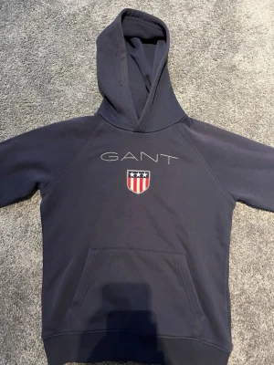 Mörkblå hoodie från GANT - Snygg mörkblå hoodie från GANT med broderad logga och sköld på bröstet. Klassisk huva och stor magficka framtill. Perfekt för en avslappnad och sportig stil. Materialet känns mjukt och bekvämt mot huden.